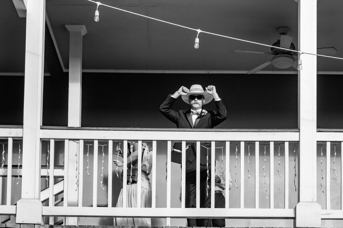 A man in a suit and hat stands on a porch adjusting his hat. The scene is in black and white.