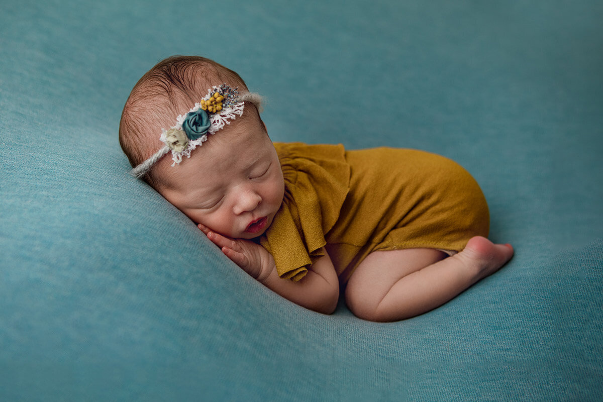 Newborn rests on teal blanket wearing mustard romper and floral headband, posed with hands tucked.