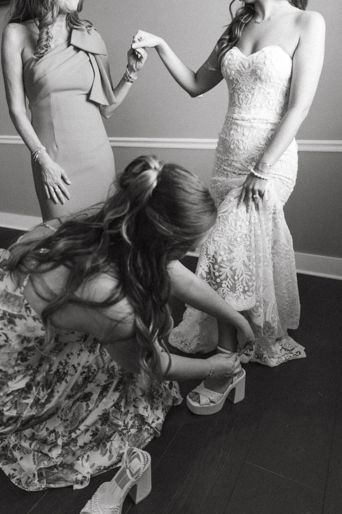 Bride and her mother holding hands while sister clasps shoe