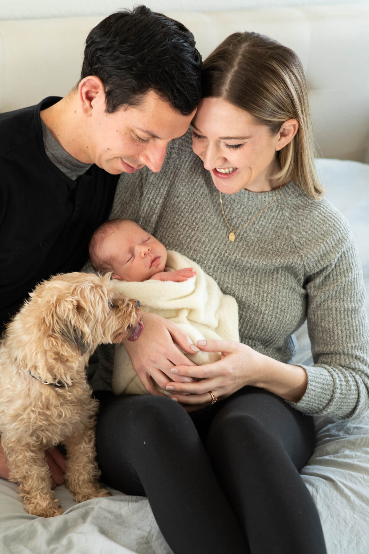 Happy new parents sit on a bed with their small dog and sleeping newborn baby between them