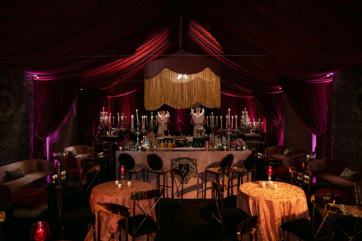 The photo was taken at a tented wedding in Virginia, captured beautifully by photographer Elisabetta Marzetti. Videographers Clayton Films were also present to document the event. The setting is a lavishly decorated event space featuring dim, romantic lighting with deep red drapes and ornate chandeliers. Tables are draped with elegant tablecloths, adorned with candles and stunning floral arrangements. In the background, two bartenders can be seen skillfully preparing drinks behind a grand central bar, adding to the elegant atmosphere of the event.