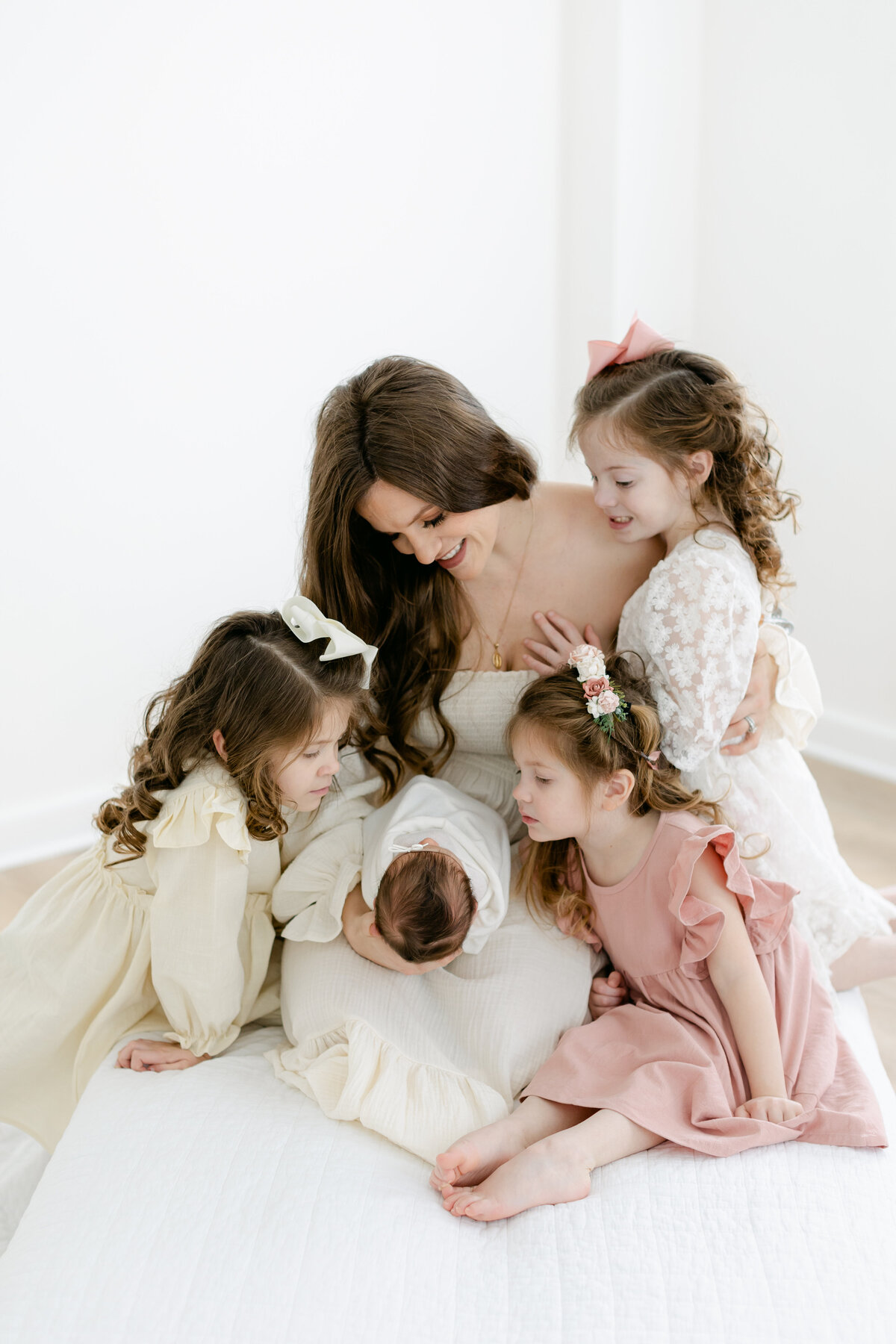 mom wearing a beautiful cream colored dress surrounded by her 3 toddler daughters on a white bed as she holds her newborn baby girl during her newborn session captured by Philadelphia Newborn Photographer Tara Federico