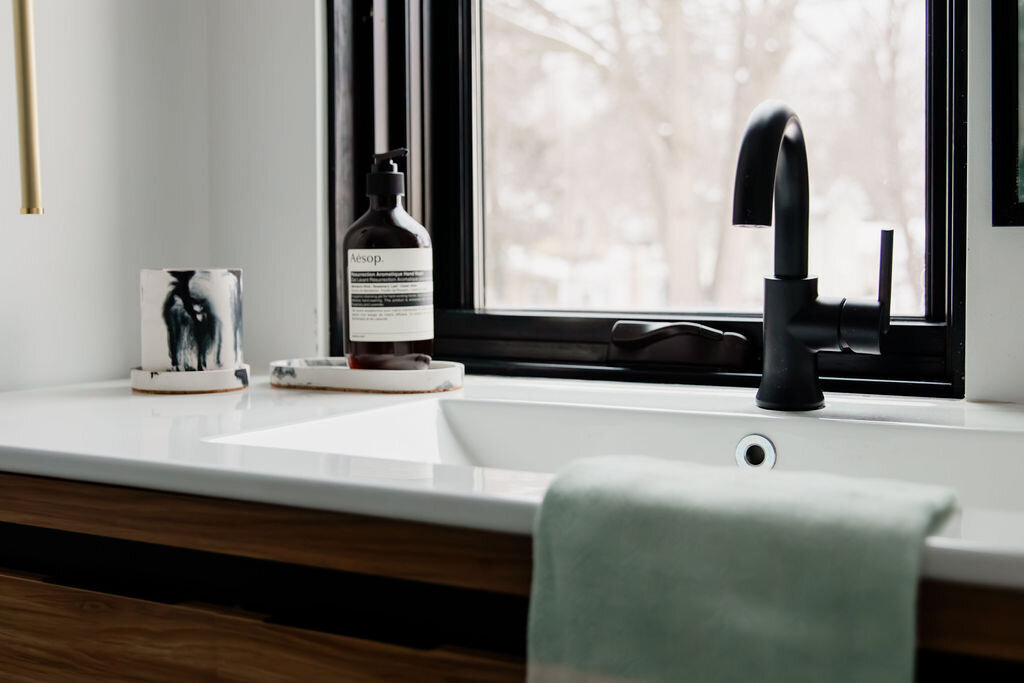 A modern bathroom sink area with a white countertop and a dark wood cabinet underneath. A sleek black gooseneck faucet is mounted on the countertop next to a rectangular white sink. A light green hand towel is draped over the edge of the sink. To the left of the faucet, a clear glass tumbler with a black and white abstract design and a black Aesop hand soap bottle sit on small, round trays. A black-framed window behind the sink offers a view of bare tree branches against a gray sky. Soft natural light illuminates the scene. A gold-toned towel bar is partially visible on the left.