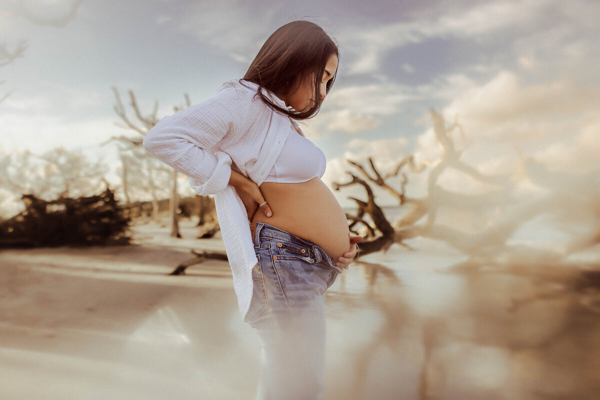 Pregnant woman in a white shirt and jeans stands on a beach with driftwood and a cloudy sky in the background.