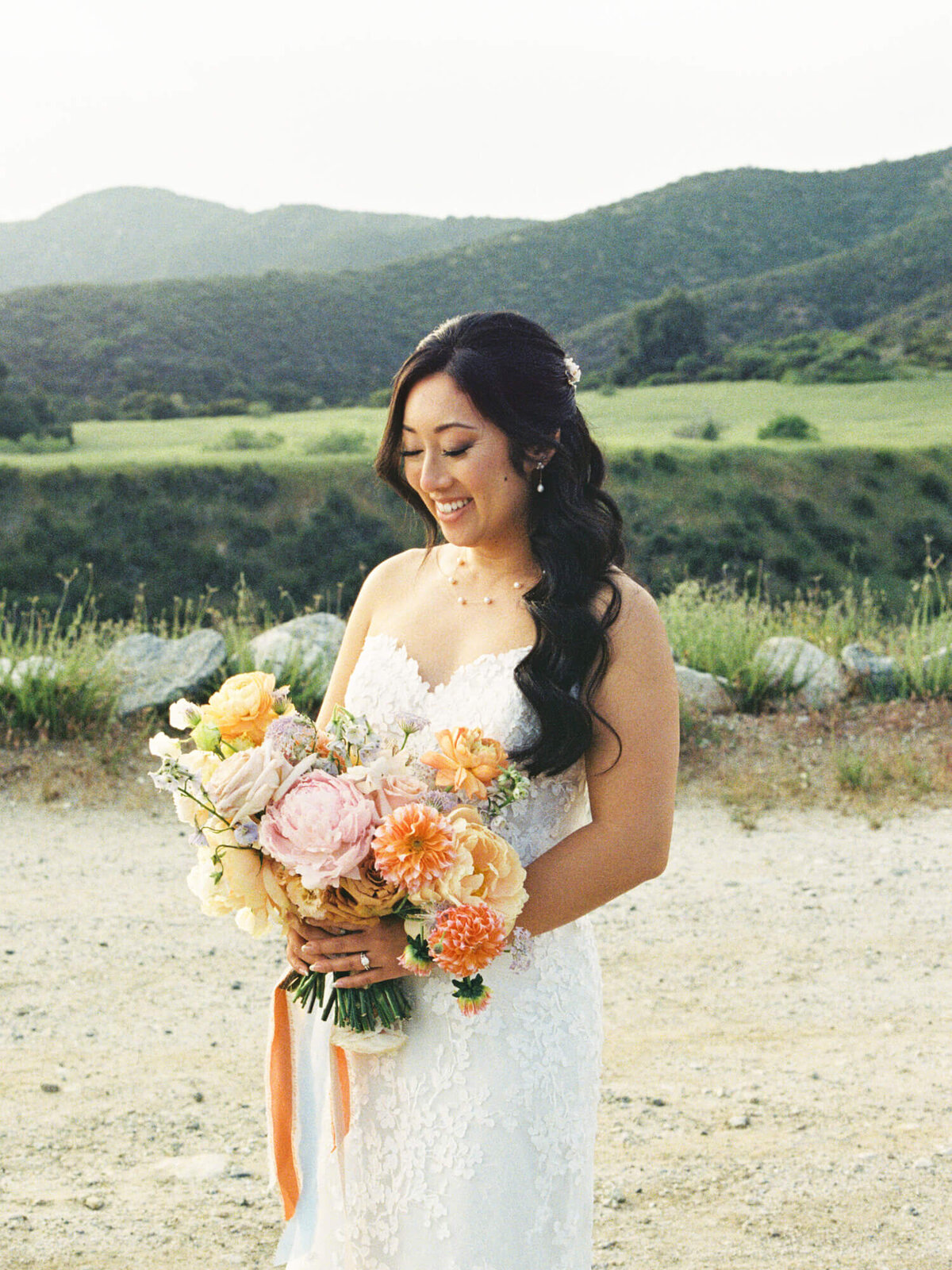 A smiling bride in a lace gown holds a vibrant bouquet of pastel flowers, set against a serene landscape with rolling hills and greenery.