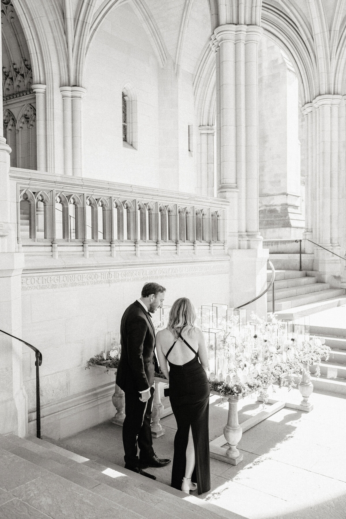 A grayscale image of a couple standing closely together at an elegant, ornate outdoor venue with grand, arched architecture. They appear to be looking at a table adorned with an arrangement of candles and flowers. The woman is wearing a backless dress.