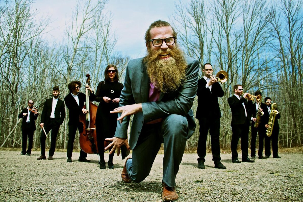 Musician portrait Ben Caplan orchestra in field with sticky trees behind them while standing in v formation Ben in front on knee