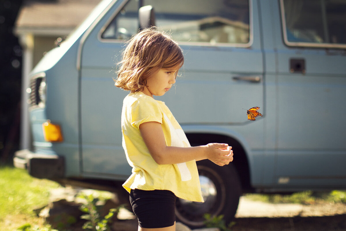 Girl in a yellow shirt with a monarch butterfly flying out of her hands. A blue Volkswagen is behind her.