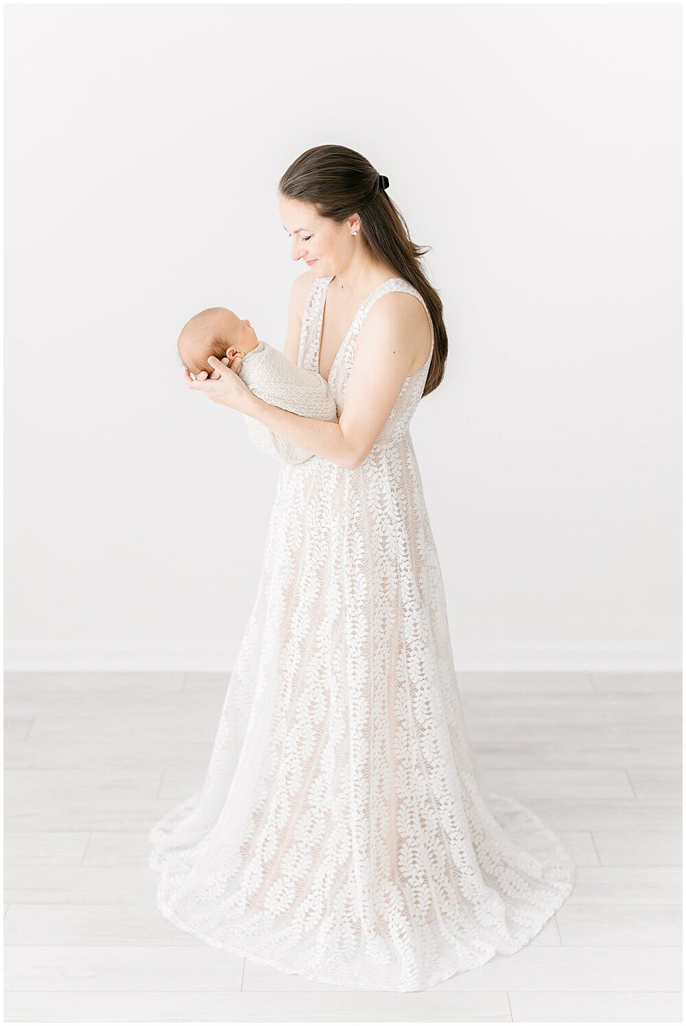 A mother wearing an elegant white and blush lace dress holding her swaddled newborn baby by washington dc newborn photographer