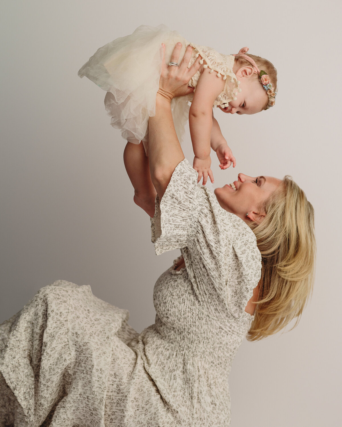 blonde mother in beautiful boho dress lifting her one year old baby up in the air while laughing