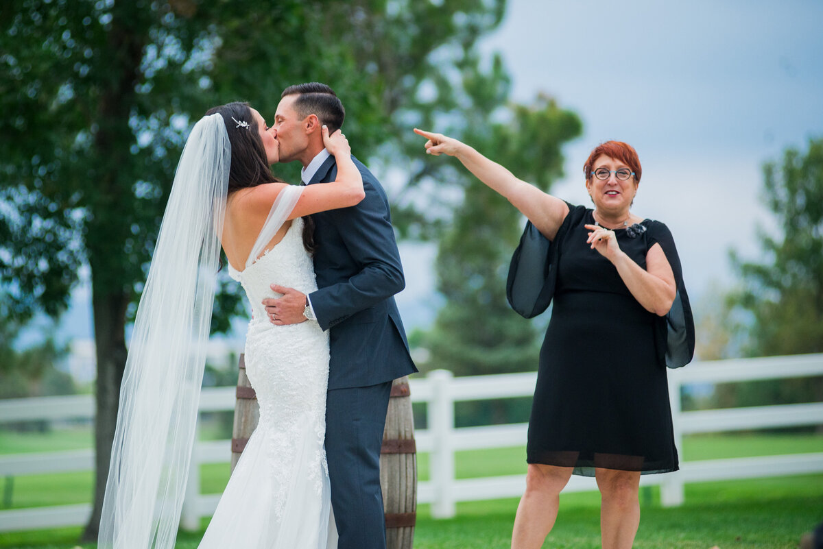 A bride and groom share a kiss as a guest jokingly stands off to the side pointing at them.