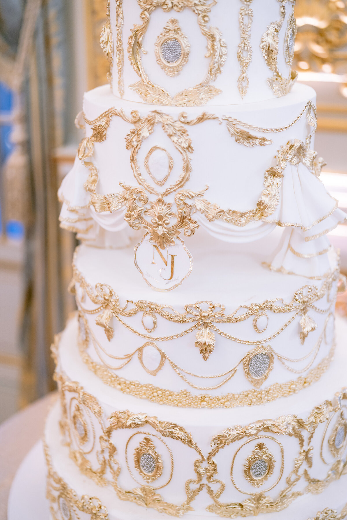 The photo was taken at the Shangri La Hotel & Ritz Paris Hotel in Paris, France by the talented photographer Matteo Coltro, with videographers Zen Film Works capturing the moment. It captures a stunning close-up of an ornate white cake, beautifully decorated with intricate gold detailing. The cake boasts multiple tiers adorned with elaborate gold lace-like patterns, delicate floral accents, and elegant garlands. A striking feature is the prominent gold letter 'J' displayed on one of the middle tiers, adding a personalized touch to the exquisite design.