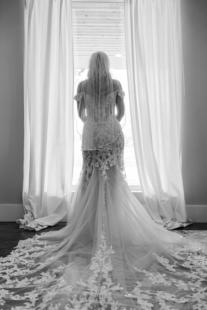 A black and white Arkansas wedding photographer image of a bride standing in a window with her long lace train spread behind her