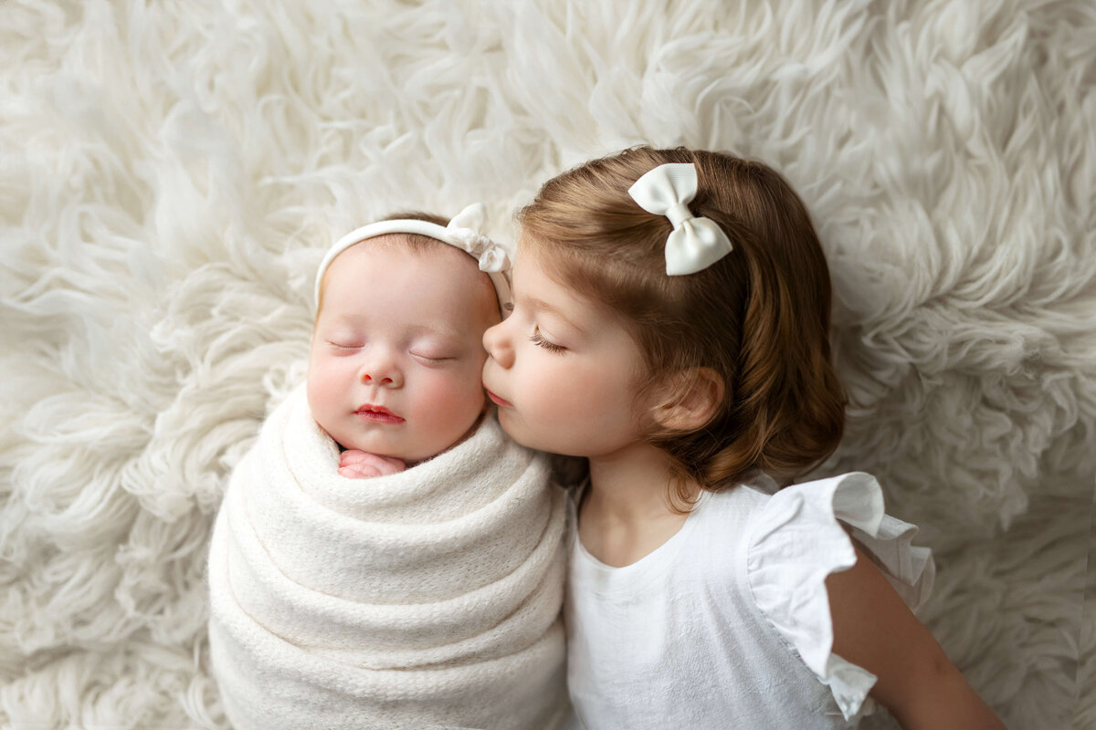 Newborn girl in a white wrap with bow laying with her big sister in a white dress kissing her at a baby photo shoot in Northern VA