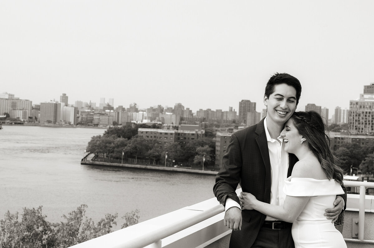 A wedding couple hugging and laughing as they lean on a railing overlooking a river 