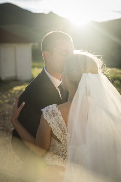 A close up moment of a groom kissing his bride on the forehead with the sun shining in the background.