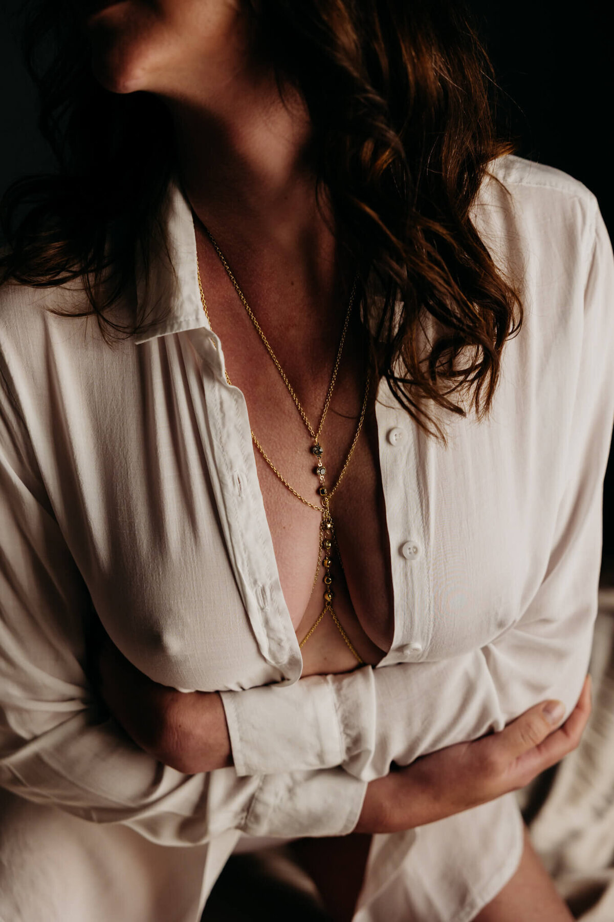 gorgeous closeup of a woman in an unbuttoned white shirt hugging herself that shows a gold chain 