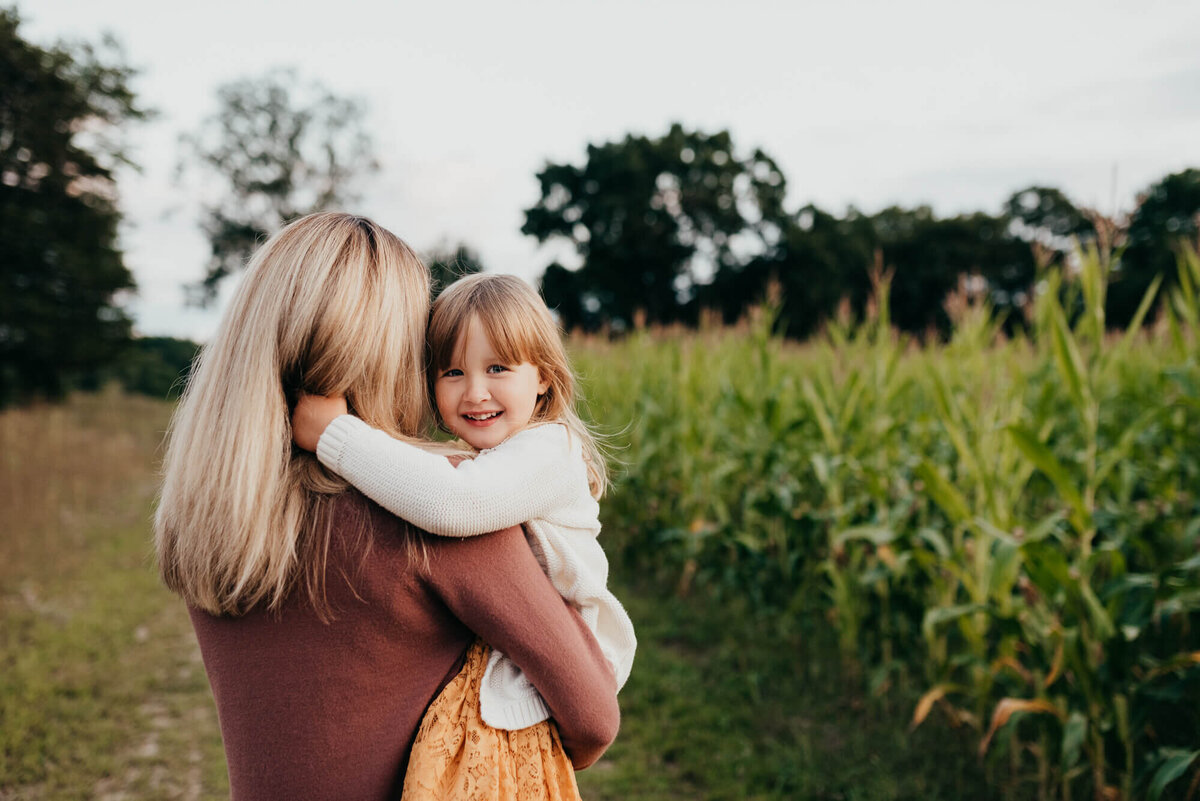 A young girl in a yellow dress and white cardigan smiles at the camera as she holds on to her mother on a path next to a corn field during their photoshoot with Allison Wolf, Boston Family Photographer