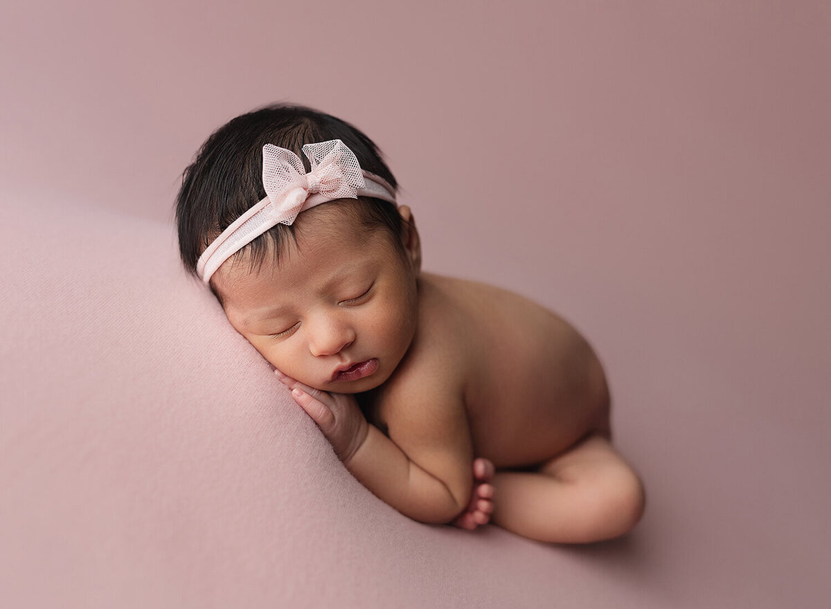A newborn baby sleeps on her belly on a pink pad with a pink bow