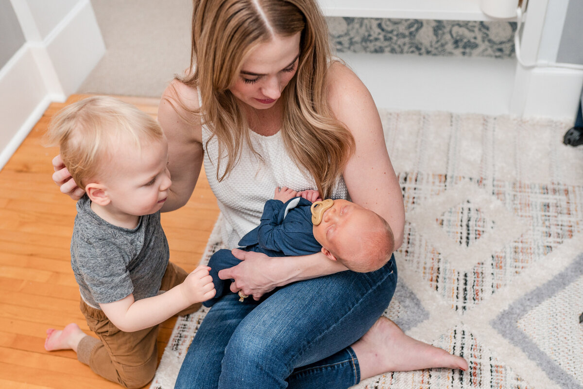 A mother sits on the floor, holding her swaddled newborn baby while her toddler stands next to them, looking down at the baby.
