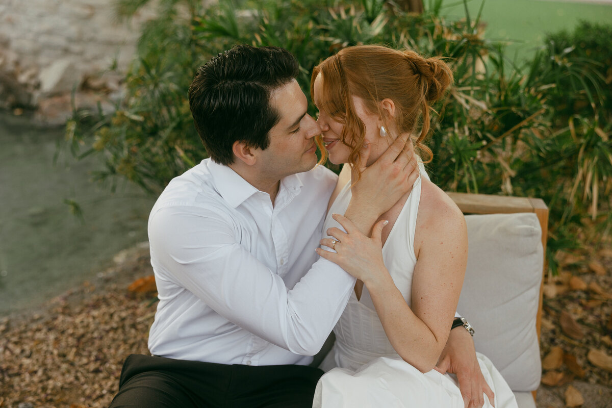 A couple sit wrapped in an embrace about to kiss, the dark haired man with his hand on the woman with red hair's face as she gently holds his arm.