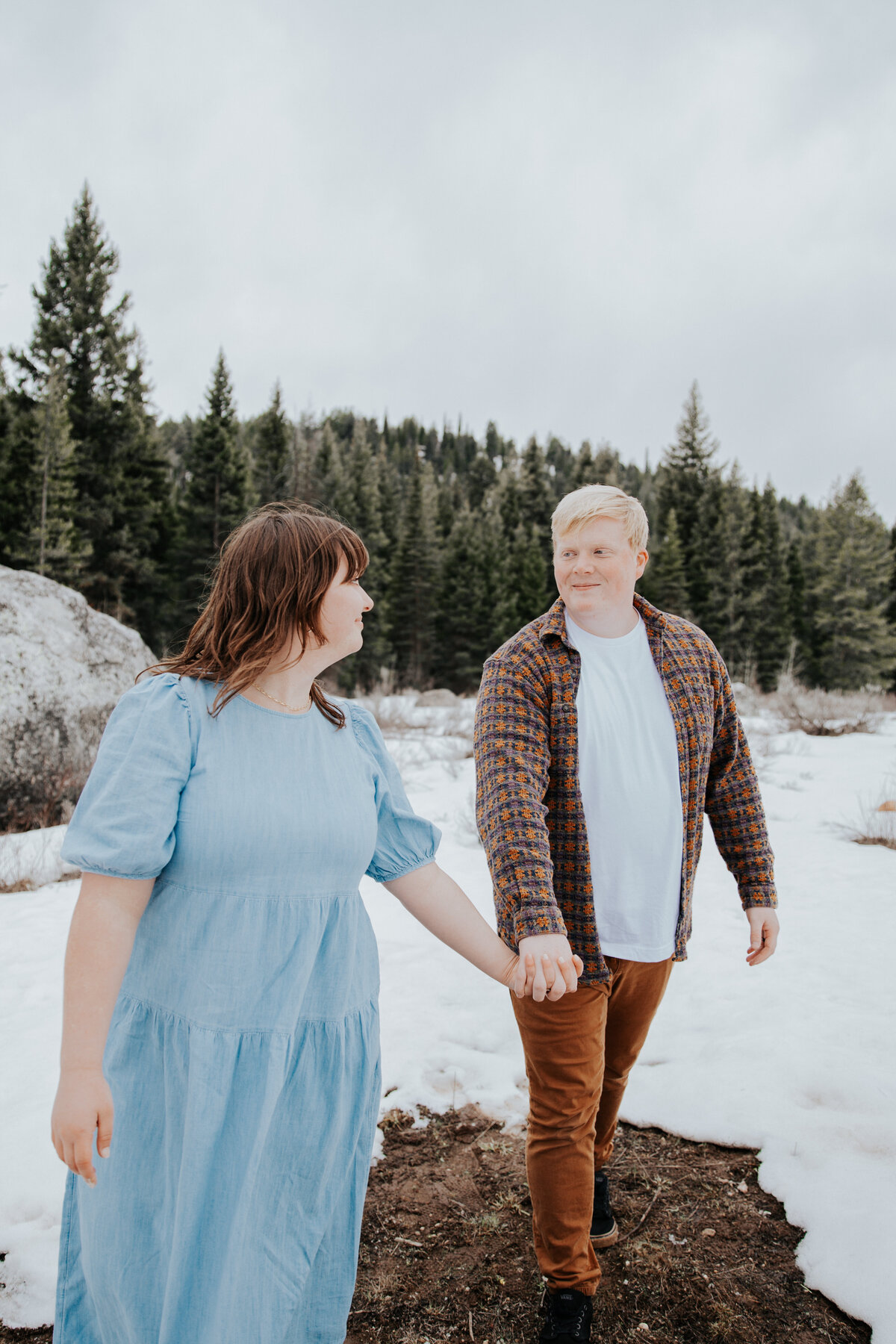 Idaho Falls wedding photographer captures engagement photos in the winter in the mountains with man and woman holding hands while walking out of the woods with snow on the ground