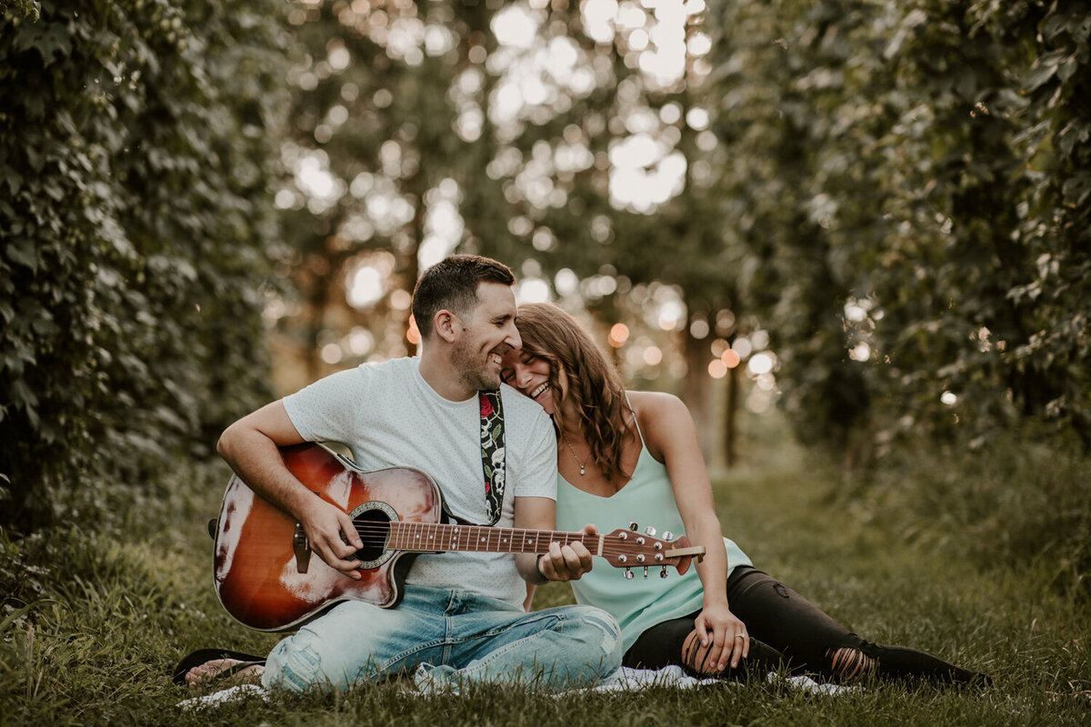 Man and woman sitting in the grass for engagement photos in Exeter, ON. The man is playing the guitar looking at the woman. The woman has her head resting on his shoulder.