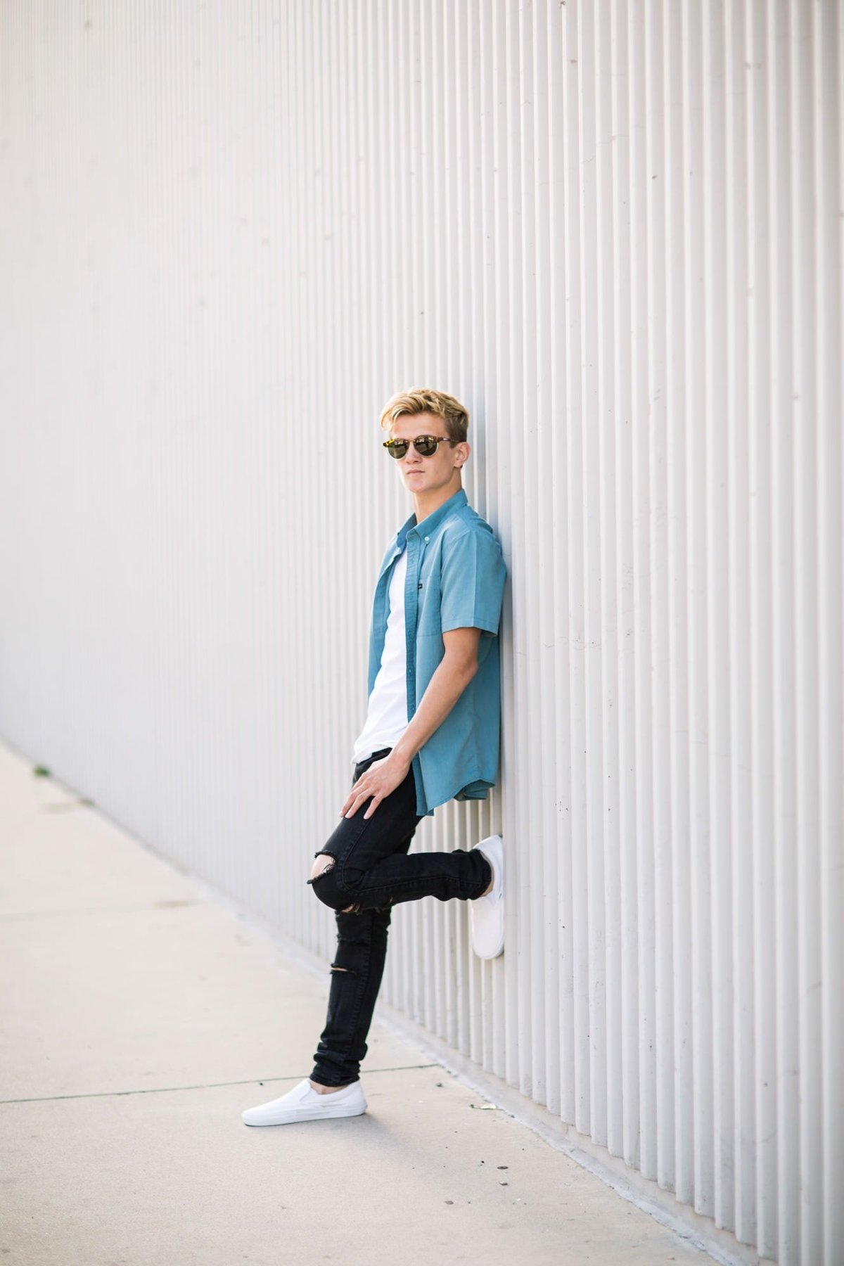 High School senior poses with sunglasses on leaning against a wall for his senior portraits