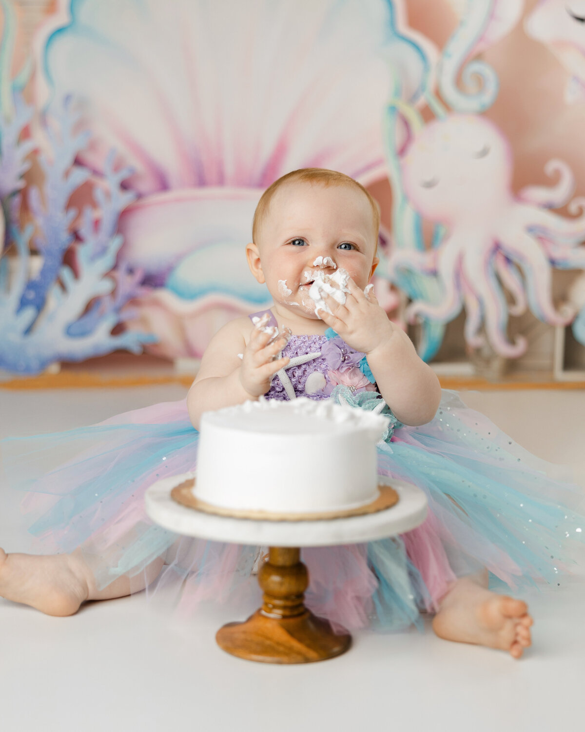 Baby in a pastel mermaid dress enjoying a cake smash with an under-the-sea backdrop. Photograph by Yucaipa cake smash photographer Kaitlyn Dawn Photography.