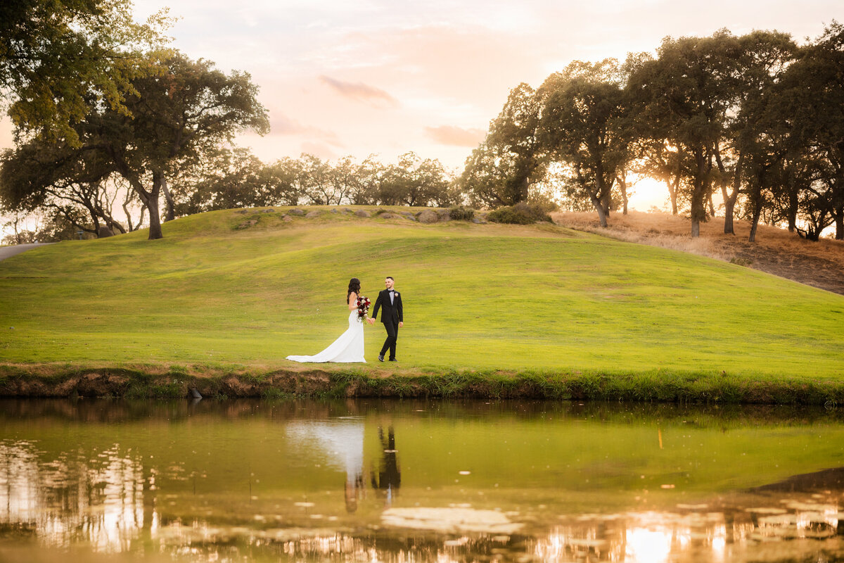 Bride and groom walk across a golf course greenery with lake in the foreground and sunset in the back.  Groom walks ahead and holds bride's hands.  Wedding photo by Sacramento wedding photography studio philippe studio pro.