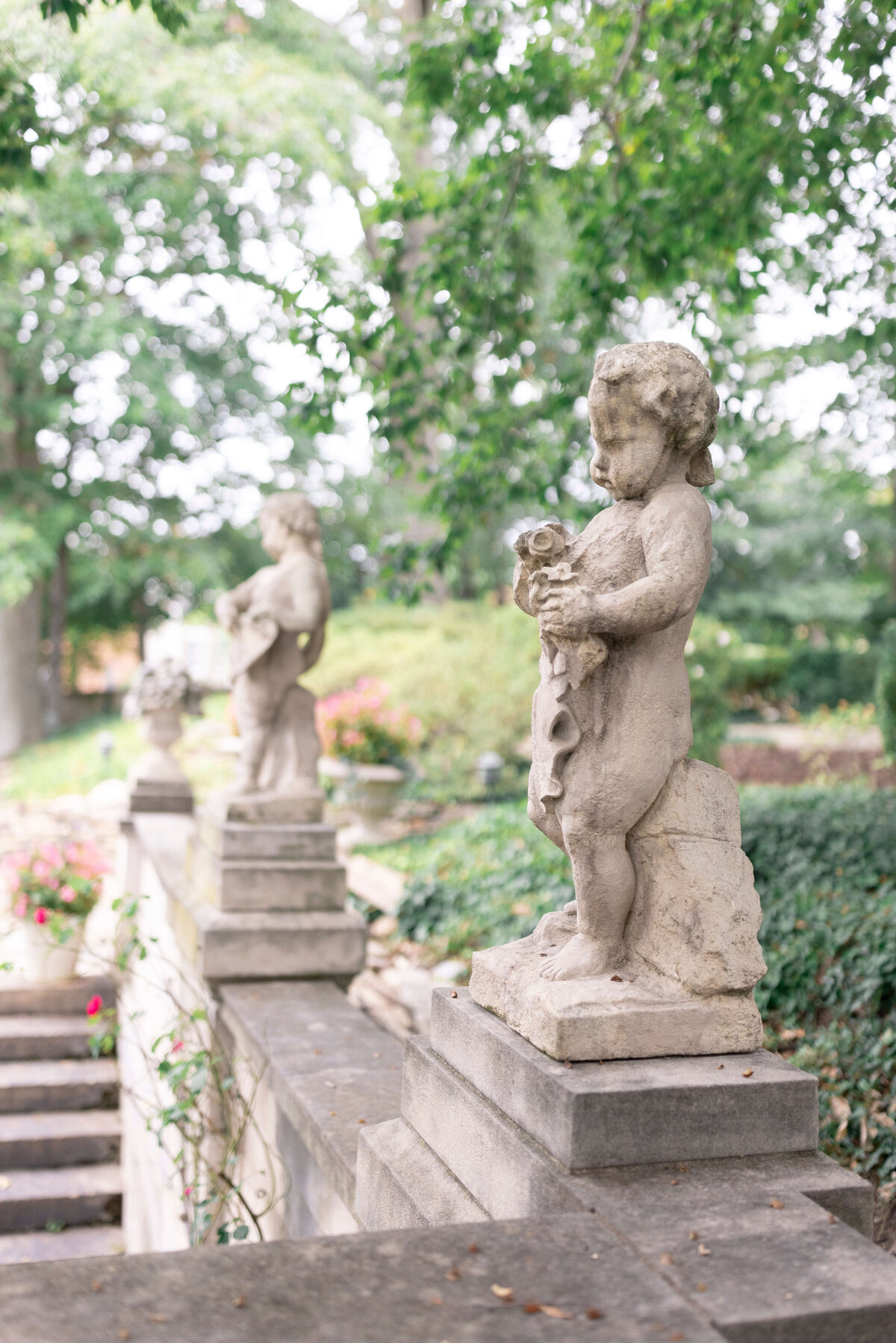 A row of cherub statues holding small animal figures adorns a garden staircase. Lush greenery surrounds the stone pathway, creating a peaceful atmosphere. The focus is on the foreground statue, with others slightly blurred in the background.