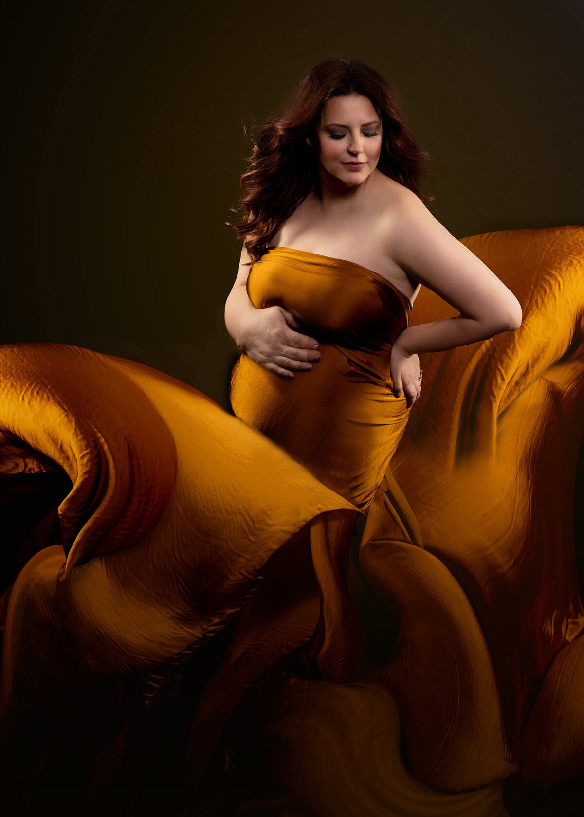 A mom to be stands in a studio with a gold sheet wrapped around her and flowing in the air all around her