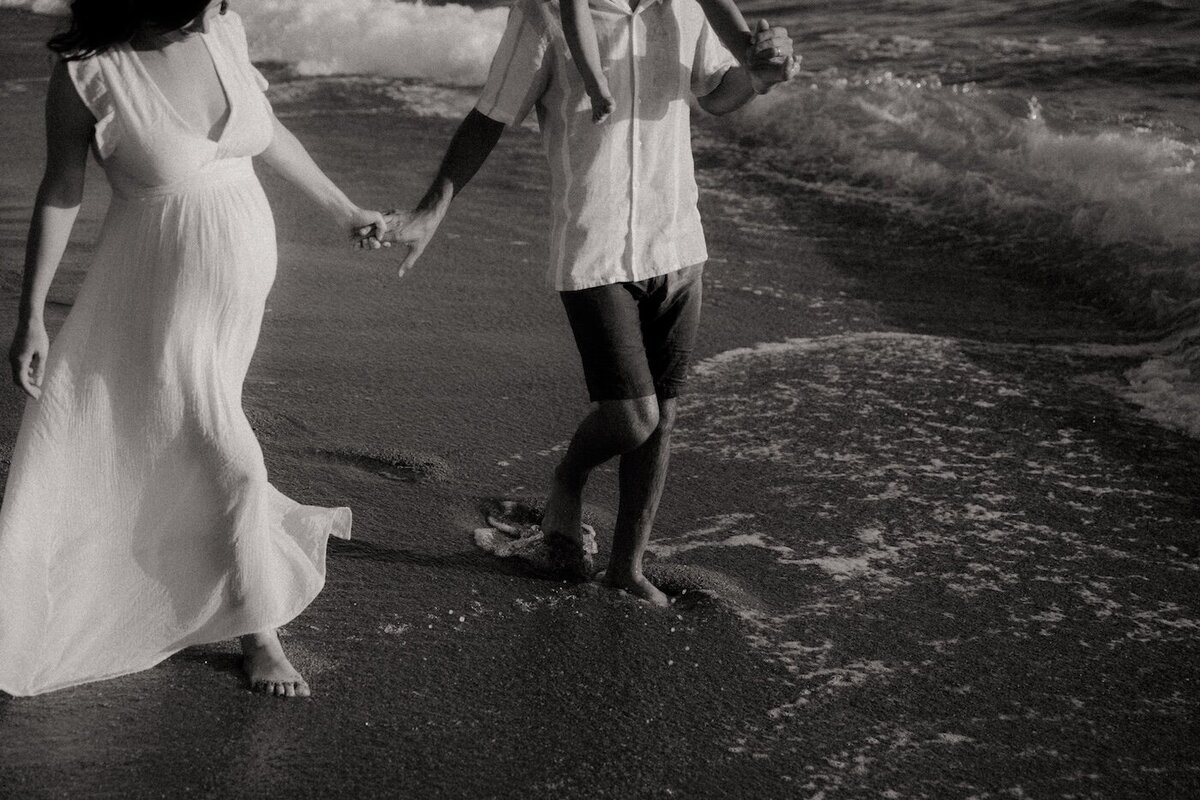 La mère enceinte marche en robe blanche, pieds dans l'eau, main dans la main avec le père qui porte leur fils. Séance photo grossesse par Kamescop, près de Nantes.