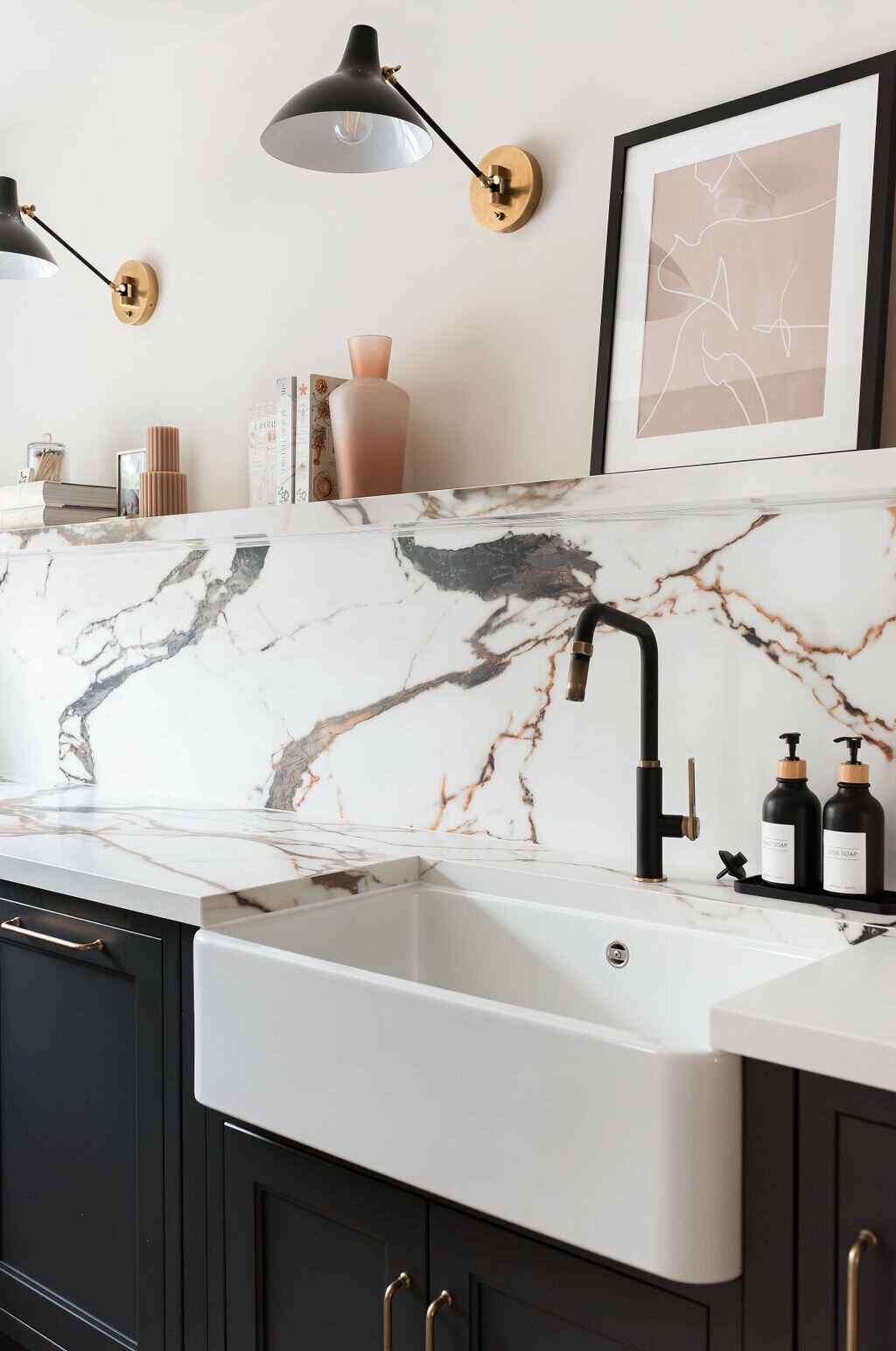 A modern kitchen with dark lower cabinets, a white marble backsplash with black and gold veining, a farmhouse sink, and minimalist open shelving holding books and decor. Two wall-mounted lights illuminate the space, which also features light wood flooring.