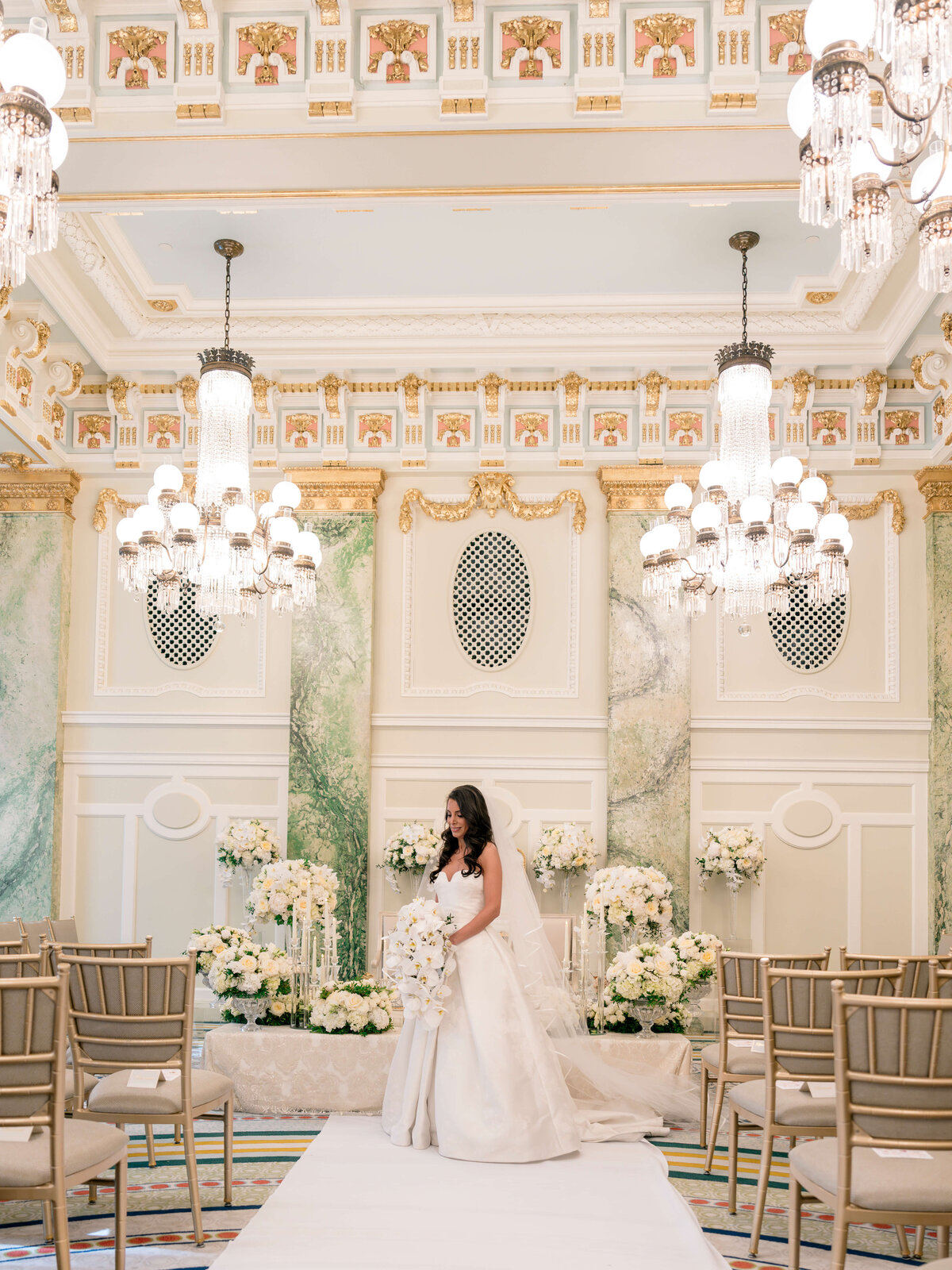 A bride in a white gown stands in an elegant, ornately decorated wedding venue. The room features crystal chandeliers, green and gold accents, and floral arrangements. Rows of empty chairs line the aisle.
