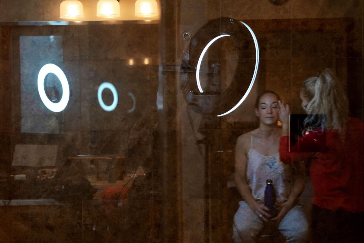 a woman gets her makeup done seen in a reflection of a shower door, surrounded by ringlights