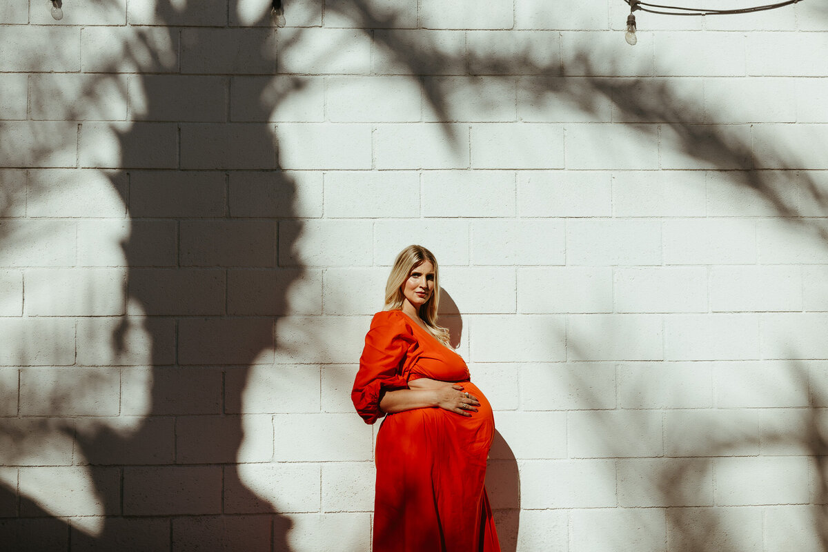Palm Springs pregnant mom poses for portrait in orange dress looking at camera with shadow of palm trees on white wall