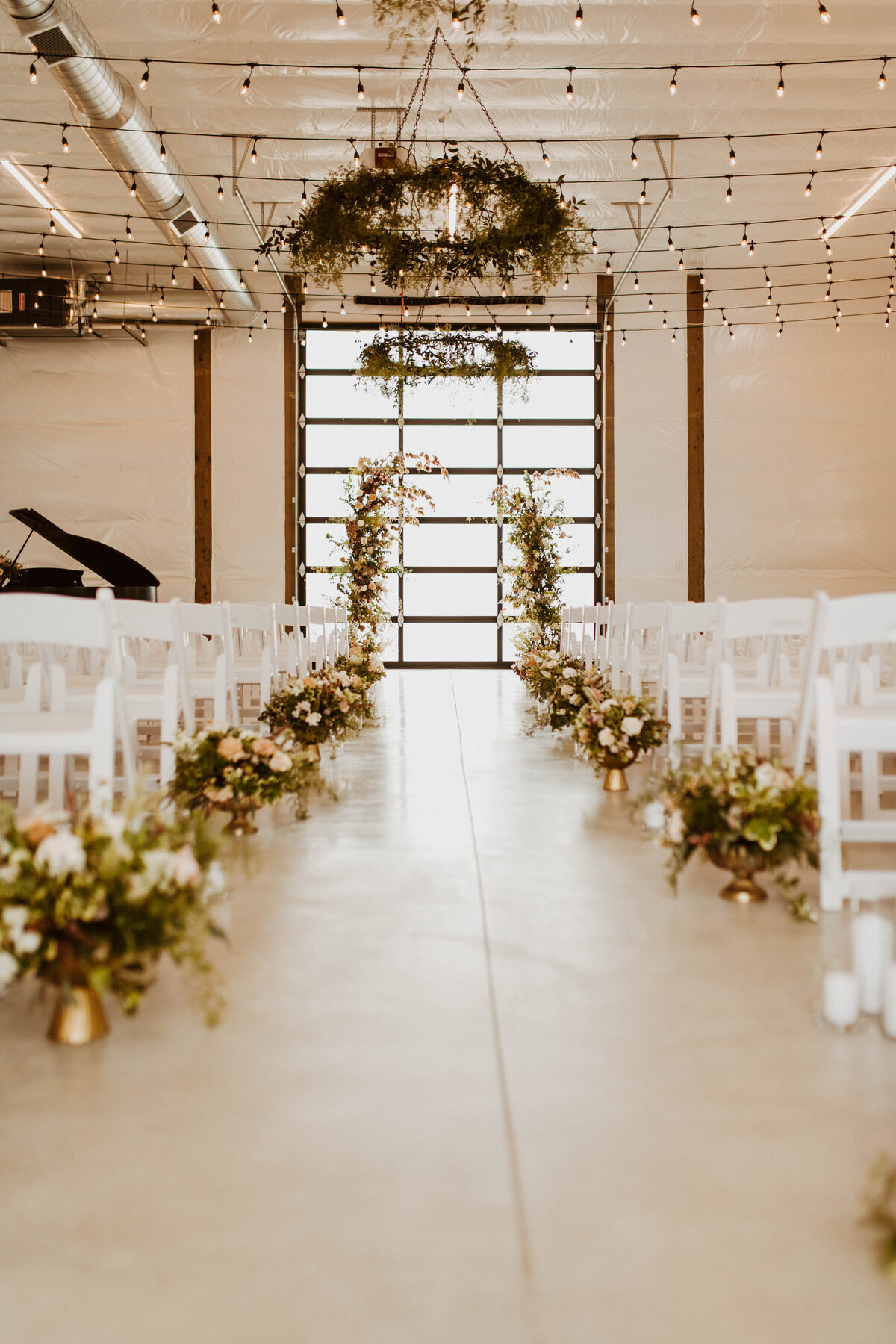 Shot of interior White Barn with rows of white chairs, flower placements at each row and a shot of the altar,  the big glass door framed with greenery and flowers.