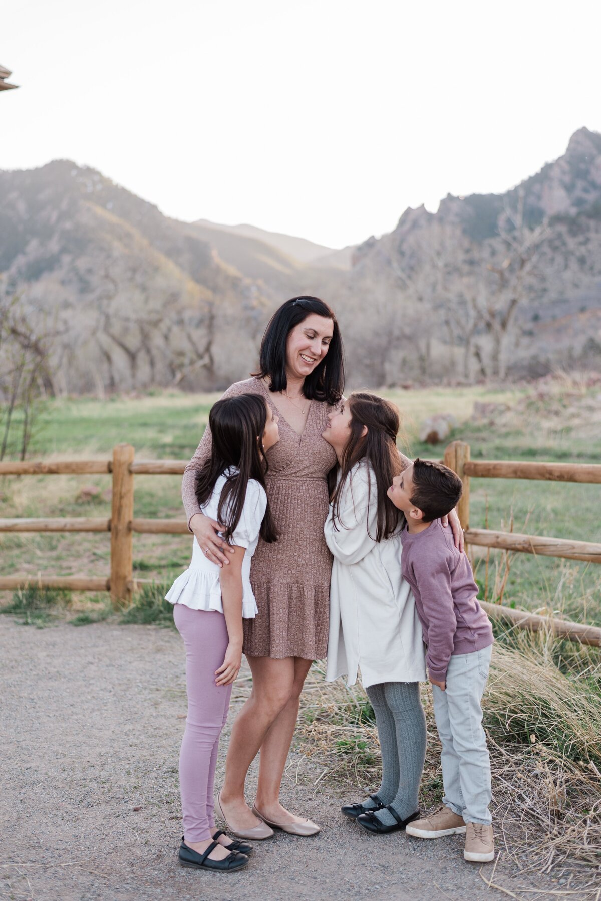 A mom smiles  at his two young daughters and young son while they hug each otherand they all look at each other captured bydenver family photographer