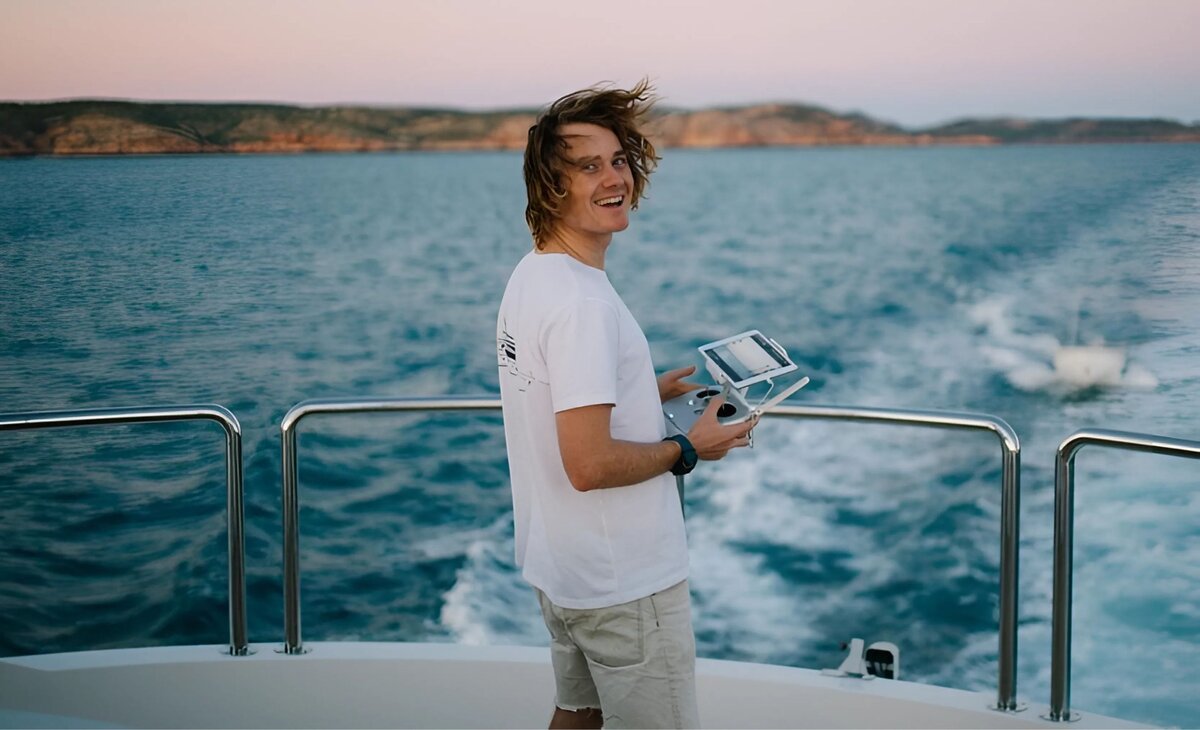 Surf style man on moving boat flying a drone and holding remote control