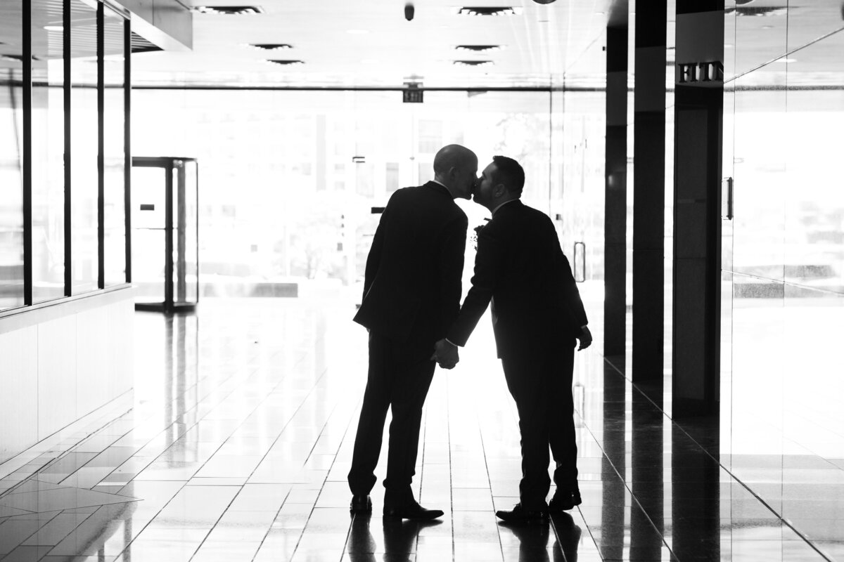 Two men share a tender moment, captured by an Austin wedding photographer, as they exchange a heartfelt kiss in a bustling hallway.