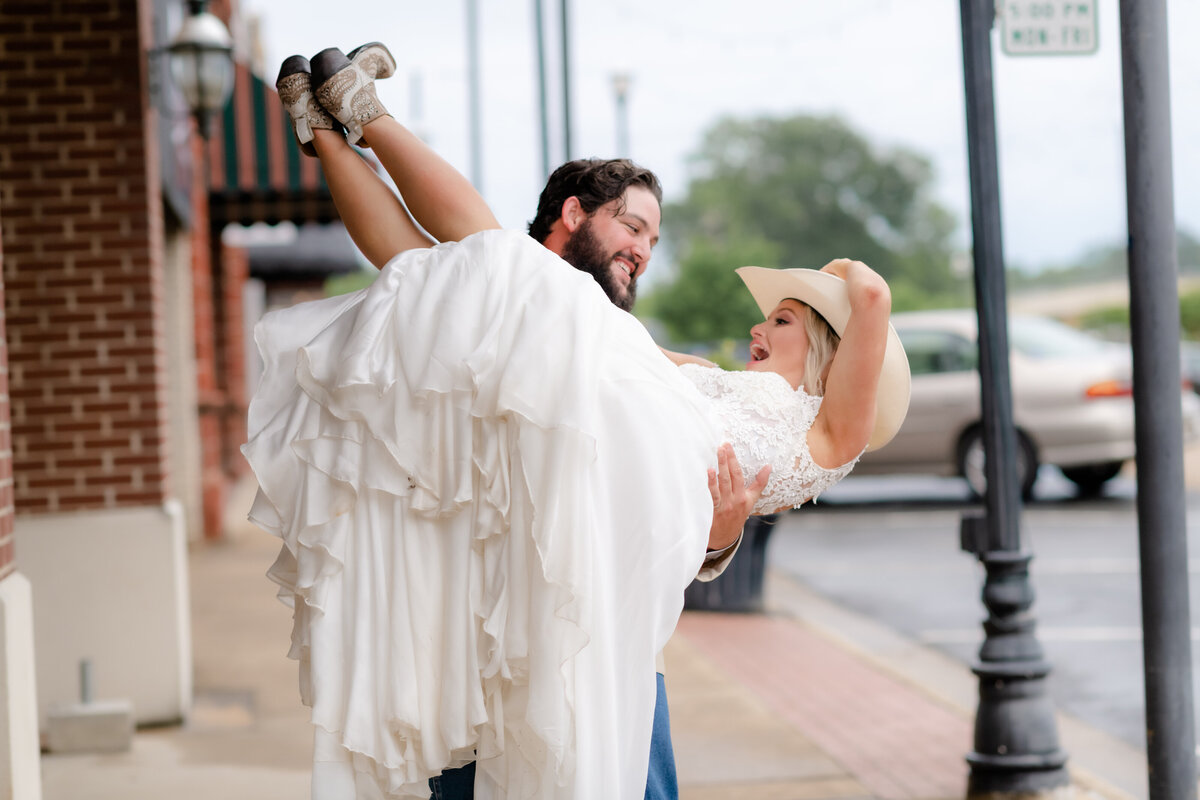 groom picks up his bride and carries her down a sidewalk as the bride wears the grooms cowboy hat