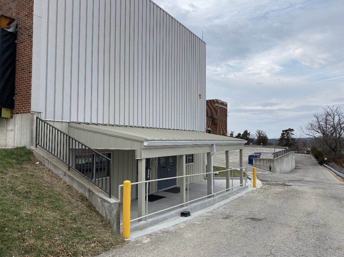 Steel awning structure with metal safety railing and yellow bollards installed at a commercial facility entrance.