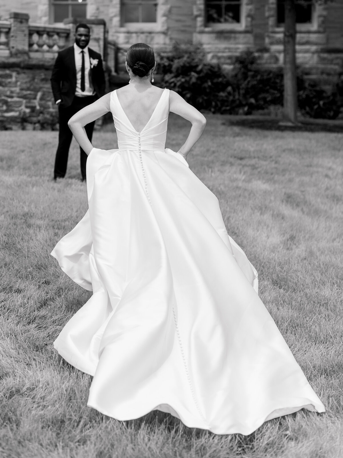 Black and white photograph of a bride walking across the lawn at Cairnwood Estate, her satin ballgown with a dramatic train and deep V-back flowing behind her. Captured by Philadelphia Fine Art Wedding Photographer Elizabeth Kane, this editorial moment reflects timeless elegance, emotional storytelling, and classic East Coast wedding style.