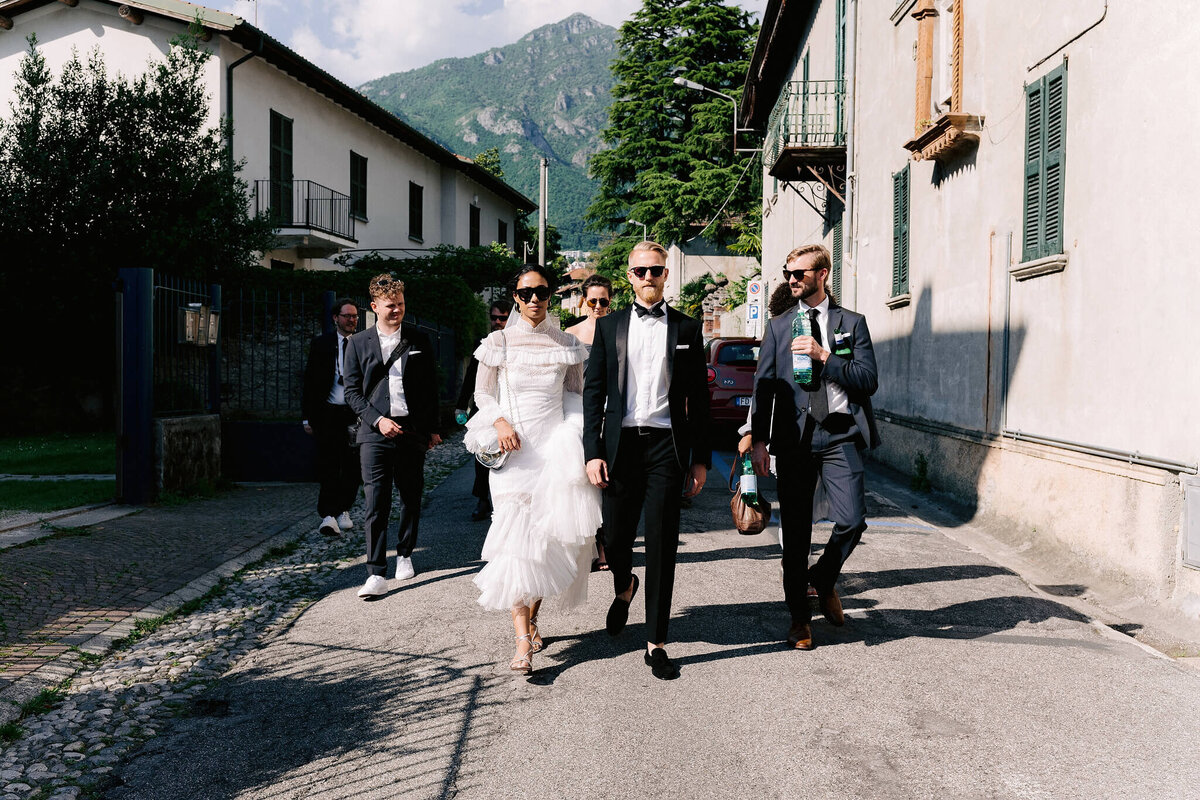 A group of people walking along with the bride and groom, in the middle of a street with houses and plants in the background