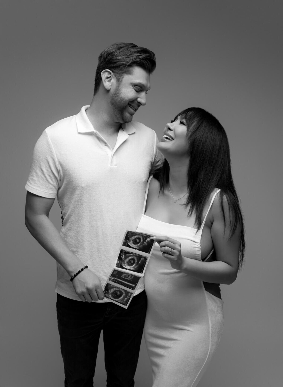 couple wearing white holding ultrasound photos and smiling at each other