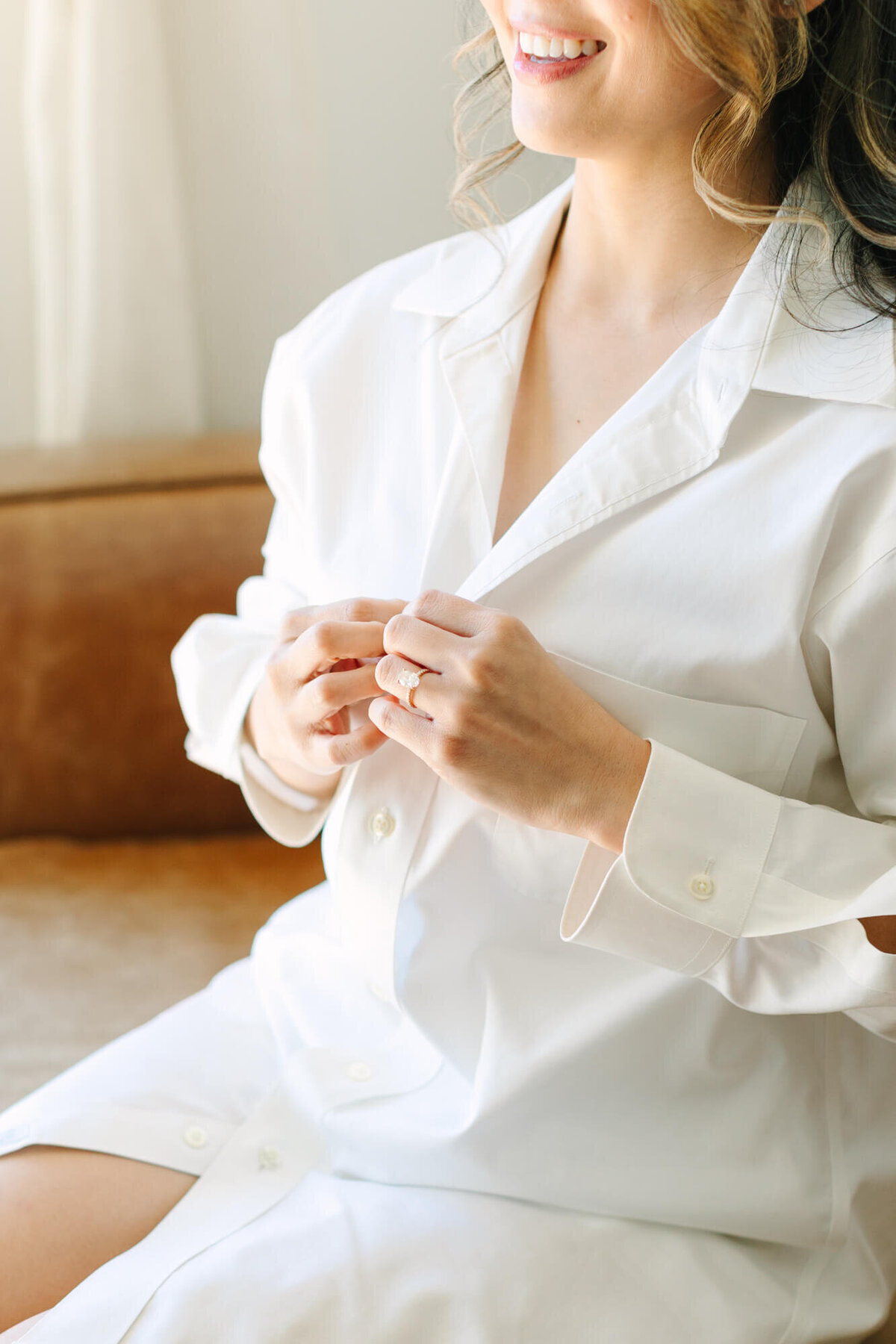 A smiling bride in a white button-up shirt sits on a brown couch, gently fastening her shirt.