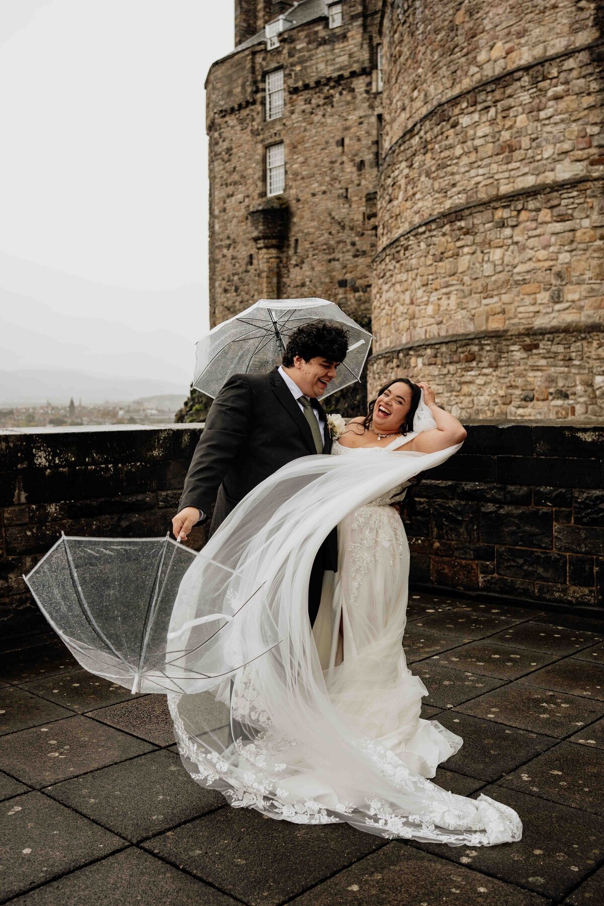 A bride and groom snuggle together as they hold umbrellas. The bride's veil blows in the wind and the laugh candidly. Edinburgh Castle is seen behind them.