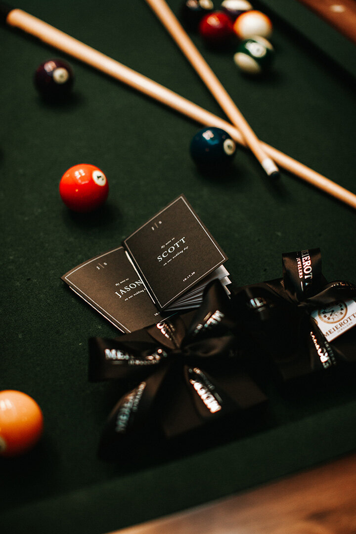 Black card stock wedding booklets with white font atop a pool table next to two black boxes with black printed ribbon.