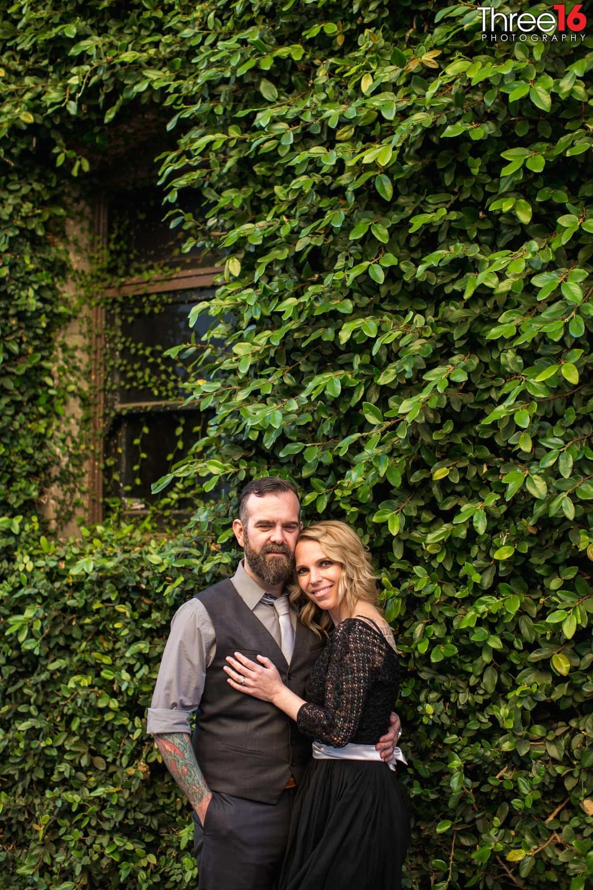 Engaged couple embrace as they pose for photos with an ivy filled wall behind them